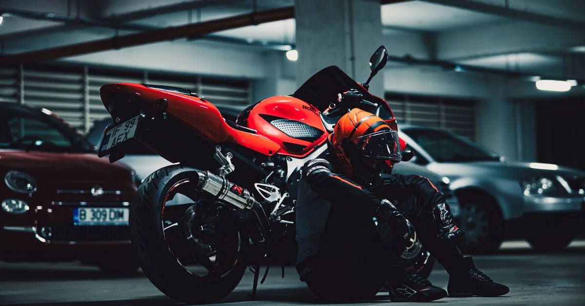Un motocycliste avec un casque est assis à côté d'une moto rouge dans un parking faiblement éclairé.