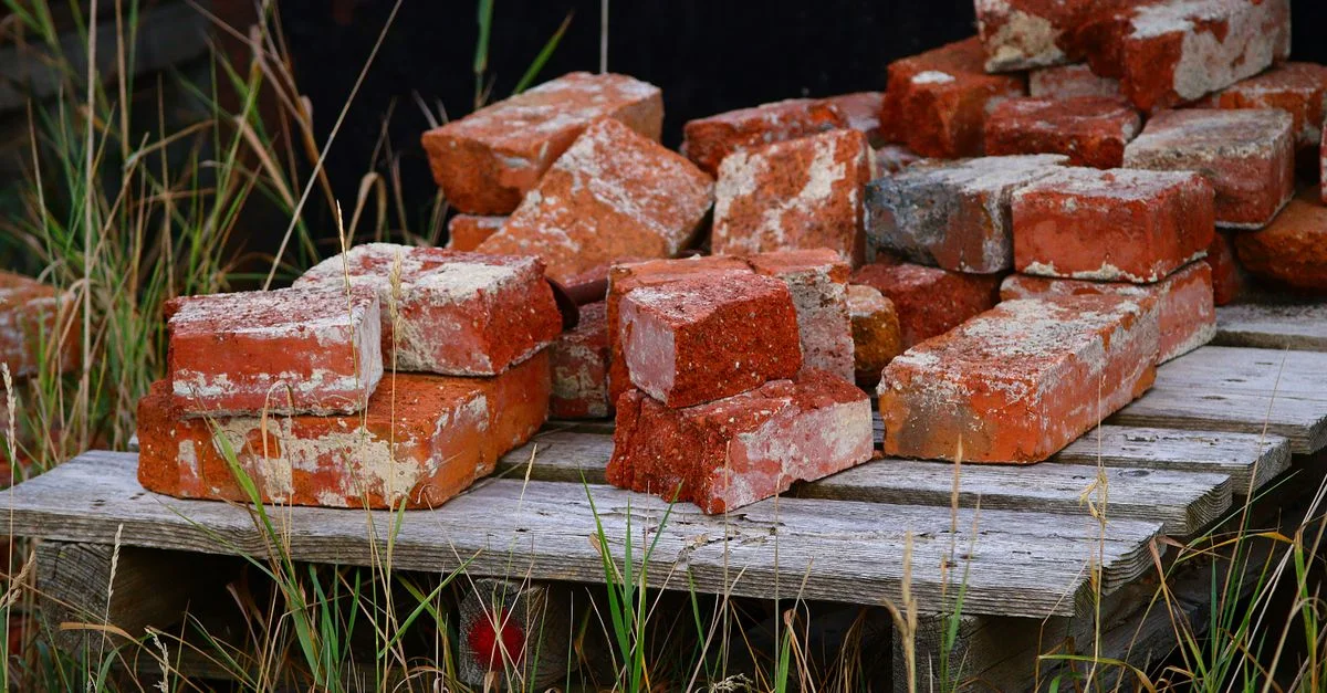 Des briques rouges patinées par le temps, empilées sur une palette en bois et entourées de hautes herbes, créent un paysage rural rustique.